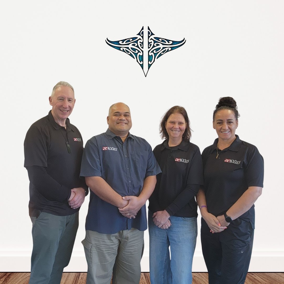 The four KYDT kaimahi in their KYDT uniforms, standing beneath the KYDT manaia